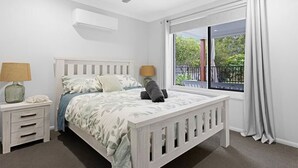 A bedroom with a bed, nightstands, a ceiling fan, a window with curtains, and a view of greenery outside.