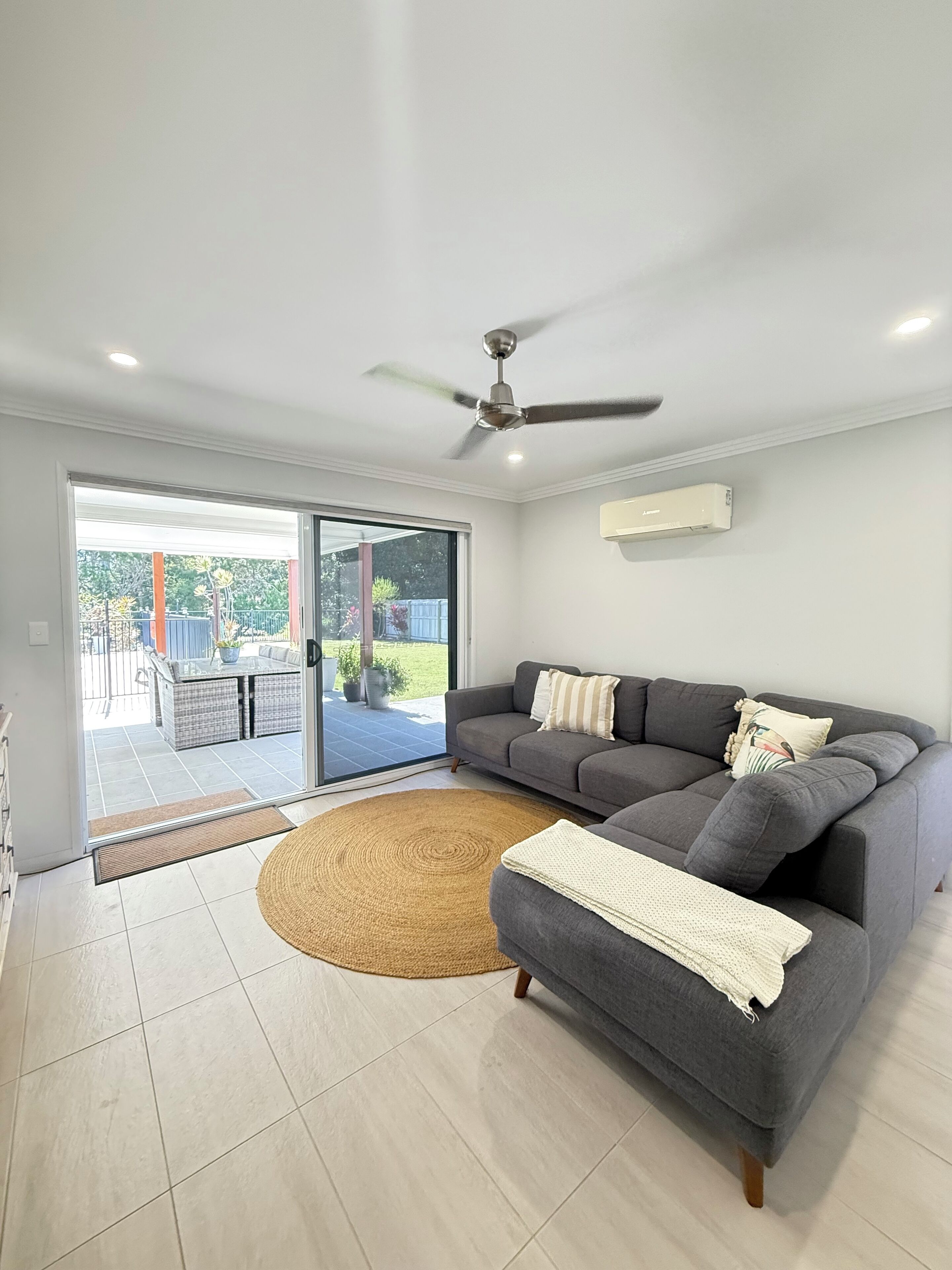 A modern living room with a grey sofa, a ceiling fan, and a view through glass doors to an outdoor patio.