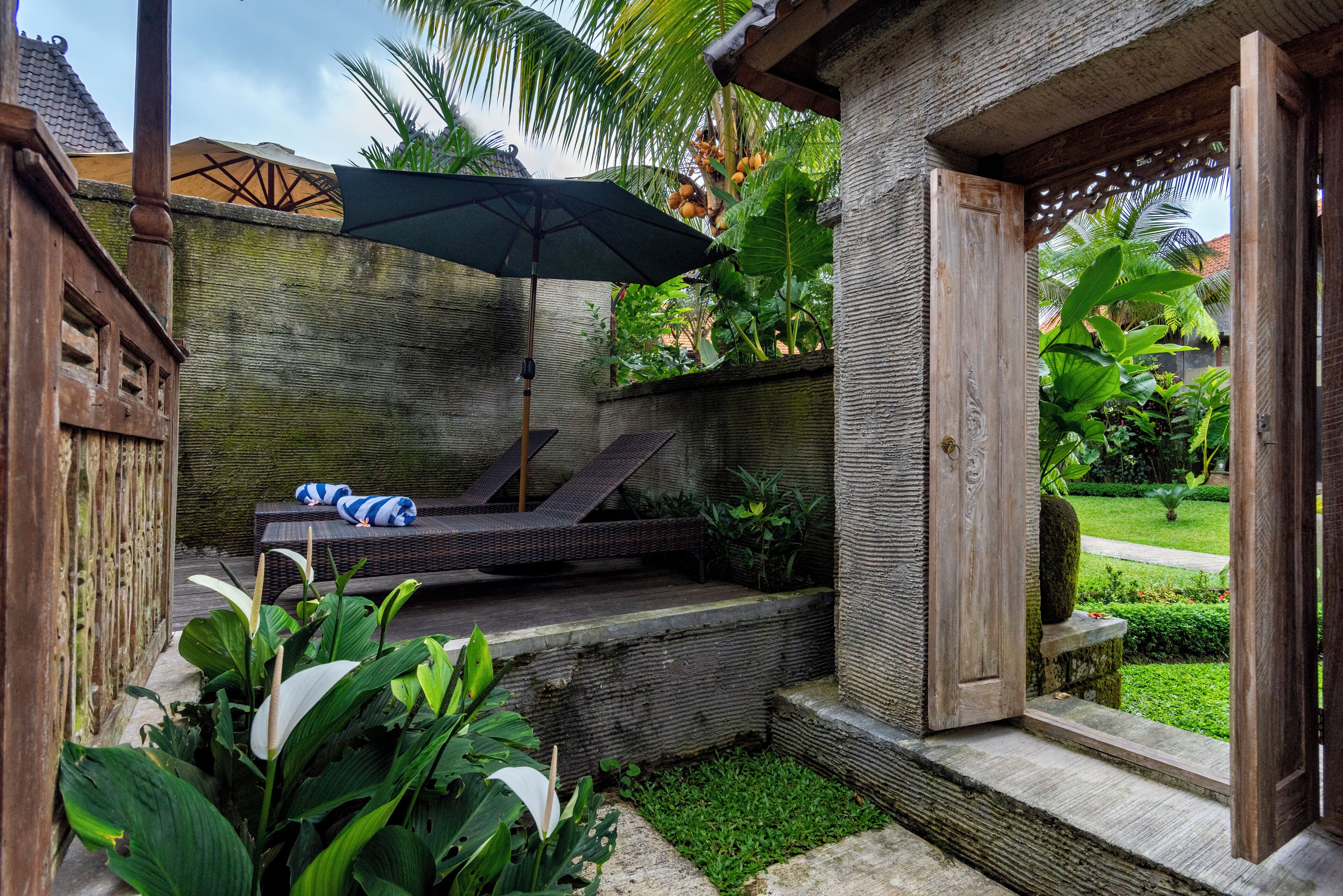 A wooden deck with a lounge chair and umbrella, surrounded by lush greenery and a stone wall.