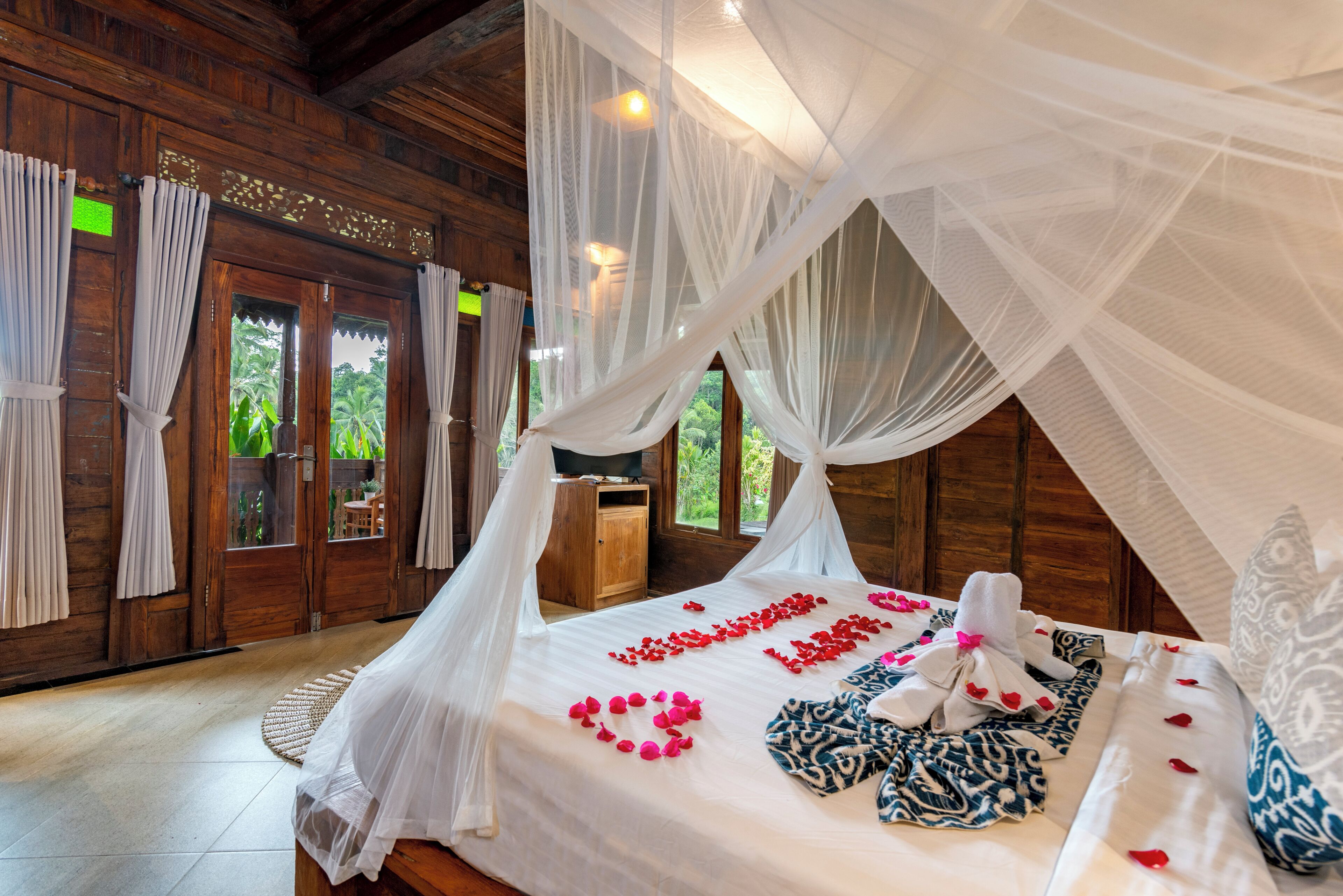 A room with a bed under a canopy, a wooden floor, and a view of greenery outside.