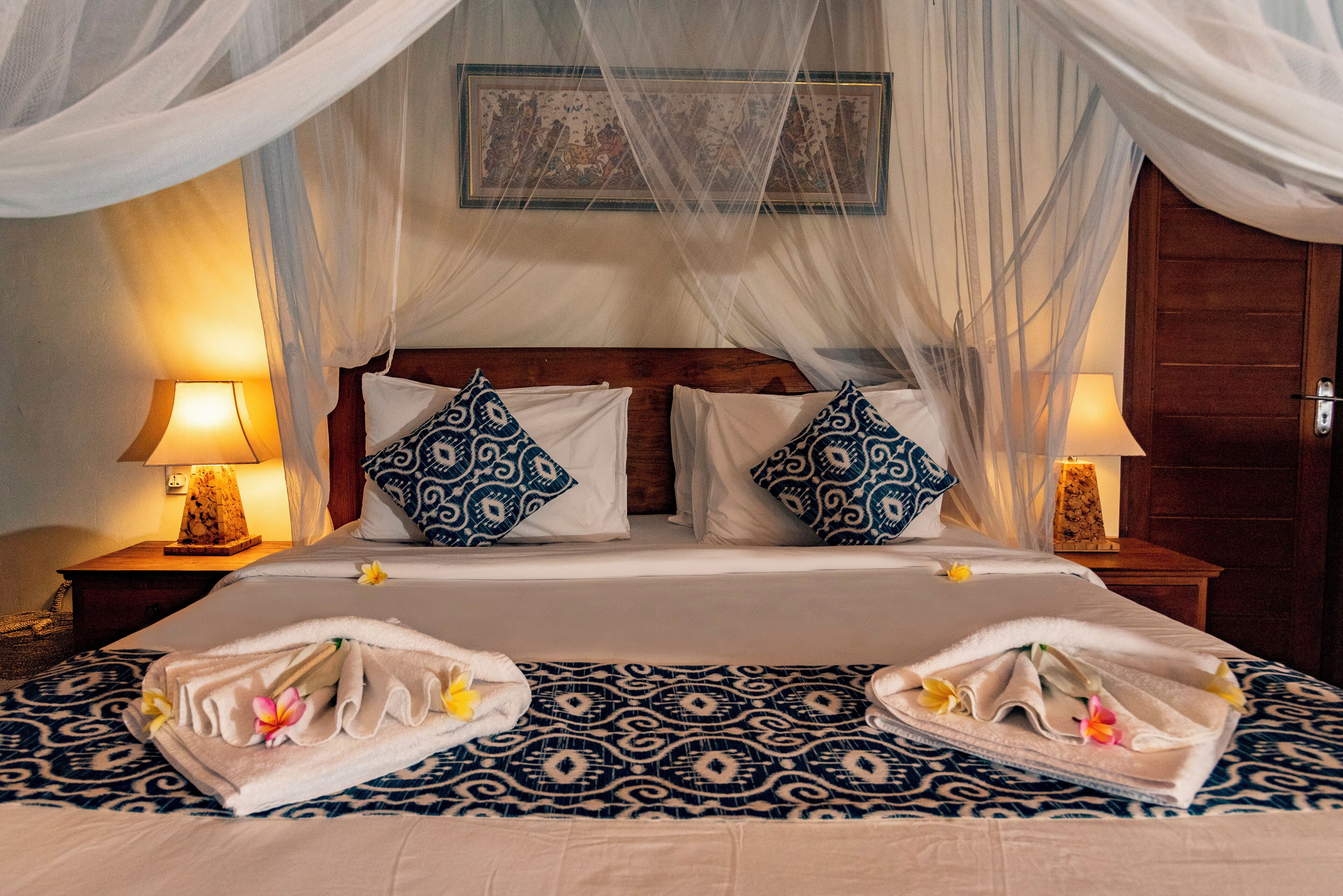 A double bed with a canopy, two nightstands with lamps, patterned bedspread, and towels with flowers.