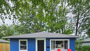 Een klein blauw huis met een zwarte deur, twee ramen en het nummer 4572 aan de zijkant. Drie rode stoelen staan er buiten.