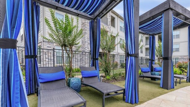 Un área de estar al aire libre con sillas acolchadas y una pequeña mesa, rodeada de cortinas azules y un edificio de piedra al fondo.