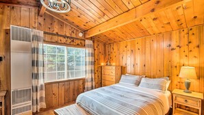 A wooden cabin bedroom with a bed, a nightstand, a lamp, a window with blinds, and an air conditioning unit. - Steps to trails • Fireplace • Pet & kid friendly (Big Bear)