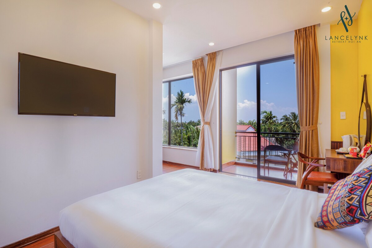 A hotel room with a large bed, a flat-screen TV mounted on the wall, a balcony with a view of palm trees, and a desk with a chair.