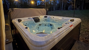 Een hottub met een deksel, omgeven door een terras, en sfeerverlichting op de achtergrond.