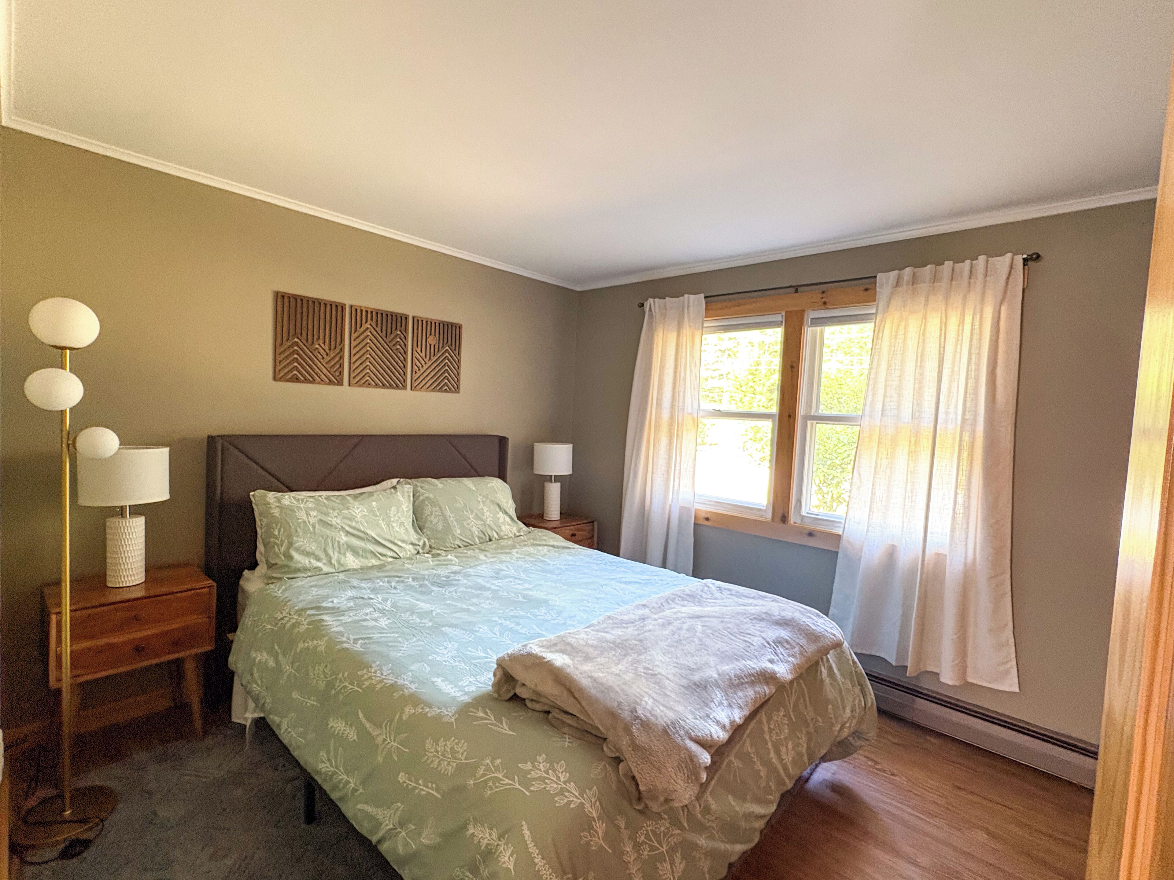 A bedroom with a bed, two bedside tables with lamps, a window with curtains, and a wall-mounted decorative panel.