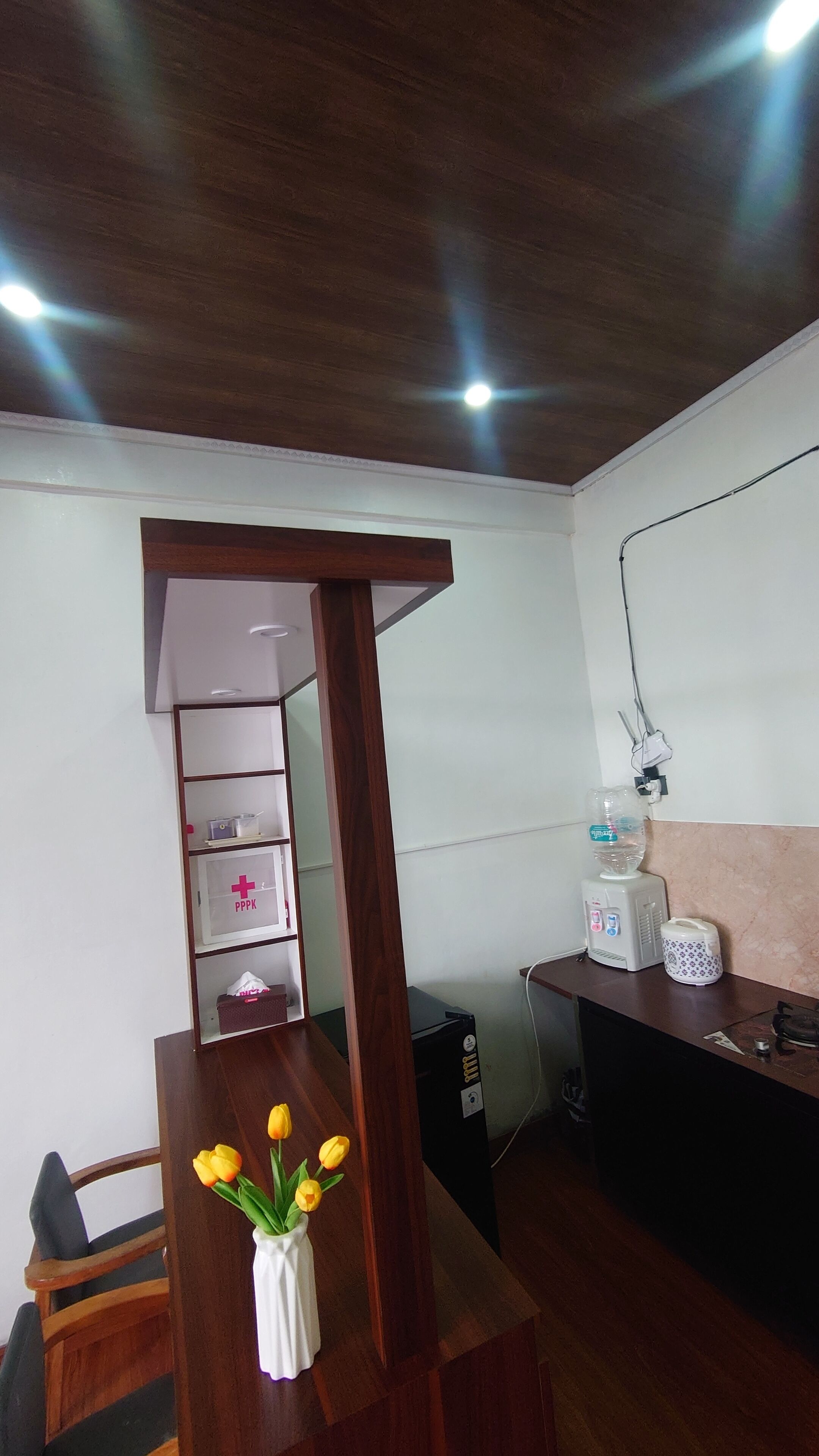 A room with a wooden ceiling, a white wall, a wooden desk with a vase of yellow flowers, a shelf with a first aid kit, and a computer on the desk.