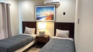 Habitación de hotel con dos camas individuales, una mesita de noche con lámpara, aire acondicionado y un cuadro con una escena de playa.