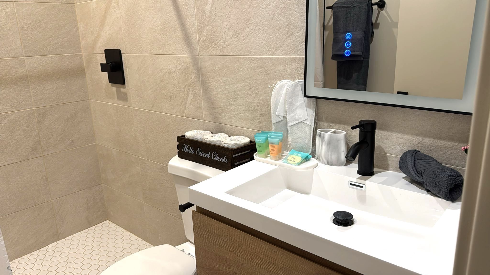 Une salle de bain moderne avec un lavabo blanc, un robinet noir et un miroir au-dessus. On y trouve un porte-serviettes avec une serviette suspendue, ainsi qu’une petite boîte sur le lavabo portant l’inscription « Bonjour, joli(e) ».