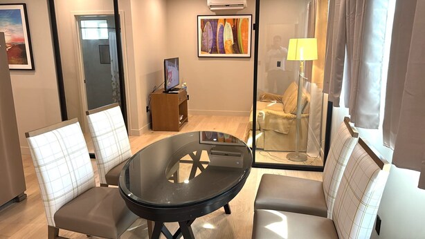 Habitación de hotel moderna con una mesa de vidrio, dos sillas acolchadas, un sofá, un pequeño armario de madera y un cuadro en la pared.
