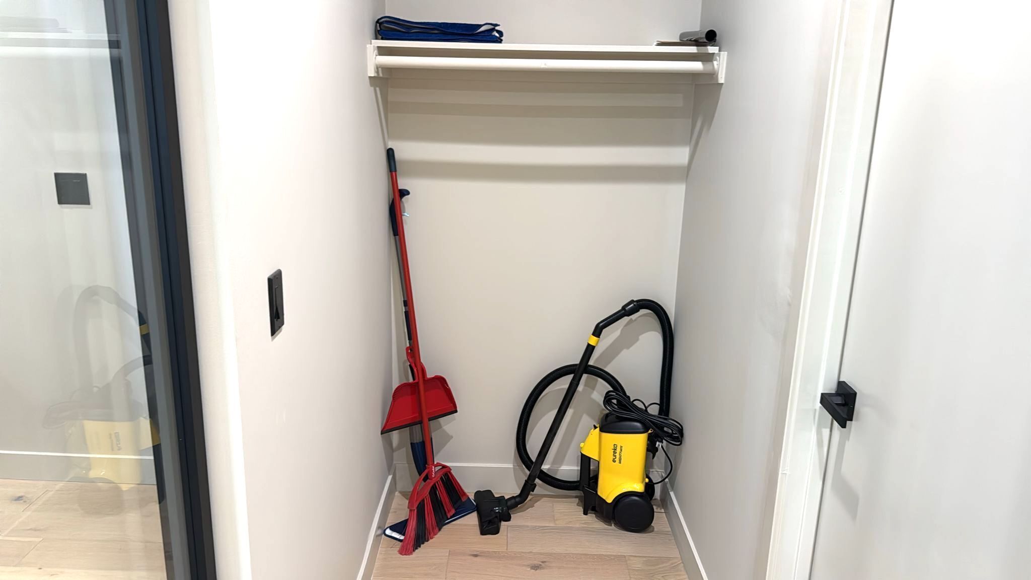 A small storage area with a vacuum cleaner, a red broom, and a blue bag on a shelf.