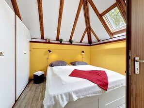 A bedroom with a sloped ceiling, wooden beams, a skylight, a bed with pillows, bedside lamps, and a small bedside table.