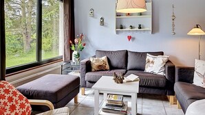 Een woonkamer met een bank, een voetenbankje en een salontafel. Er is een raam met uitzicht op bomen, een hanglamp en een plank met decoratieve objecten.