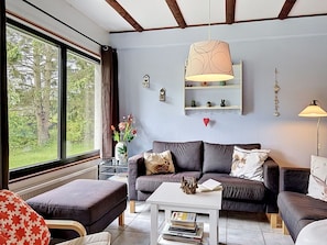 A living room with a sofa, ottoman, and a coffee table. There is a window with a view of trees, a hanging lamp, and a shelf with decorative items.
