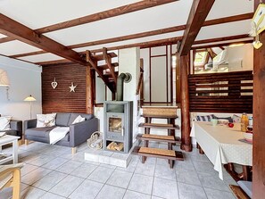 A cozy living room with a fireplace, wooden beams, a sofa, a dining table, and a staircase.