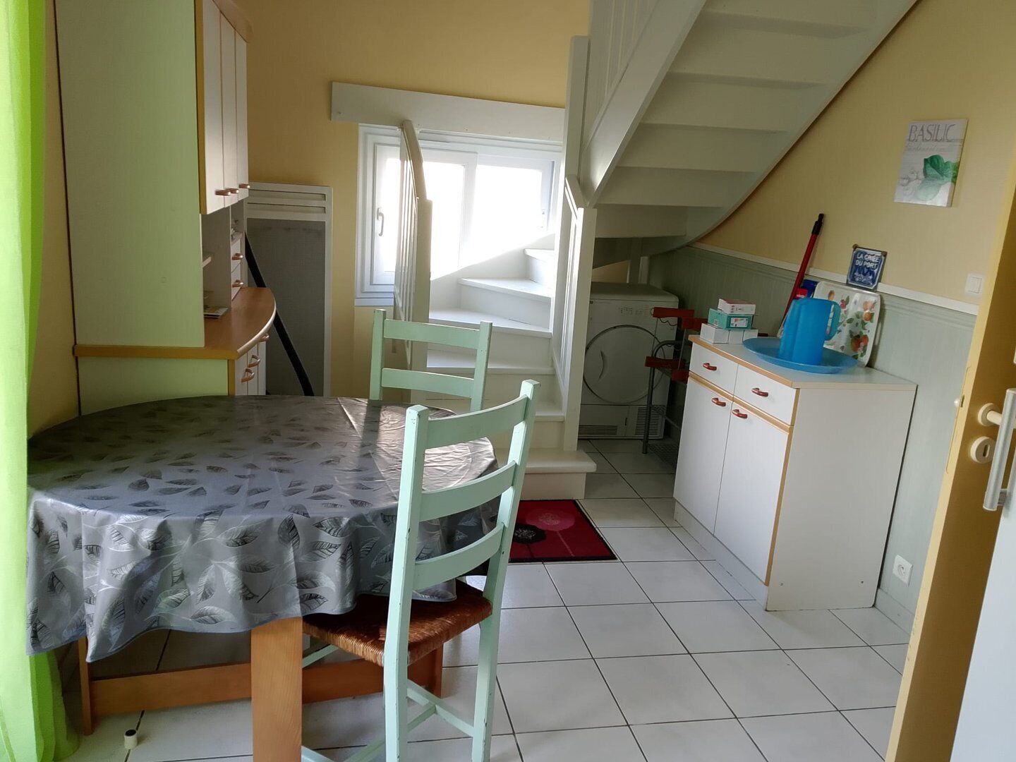 Une cuisine compacte avec un coin repas, comprenant une table recouverte d’une nappe à motifs de feuilles, un lave-linge et un petit meuble contenant divers objets.