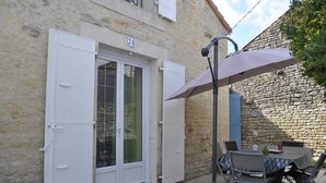 Edificio de piedra con puerta blanca marcada '24', un patio con mesa y sillas, y una sombrilla grande que proporciona sombra.