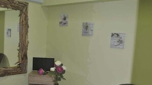 Una habitación con sofá blanco, una mesa de centro de vidrio con un jarrón de flores, una silla de mimbre, un espejo grande y una cama con cabecera.