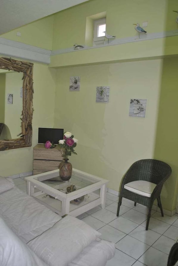 A room with a white sofa, a glass coffee table with a vase of flowers, a wicker chair, a large mirror, and a bed with a headboard.