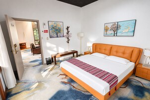 Una habitación de hotel con una cama grande, un banco, una mesita de noche, una lámpara y dos cuadros decorativos.