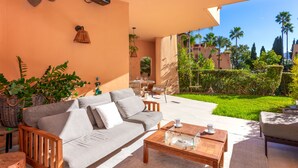 Ein gemütlicher Sitzbereich im Freien mit einer Couch, einem Couchtisch und Stühlen, mit Blick auf einen gepflegten Garten mit Palmen und blauem Himmel.