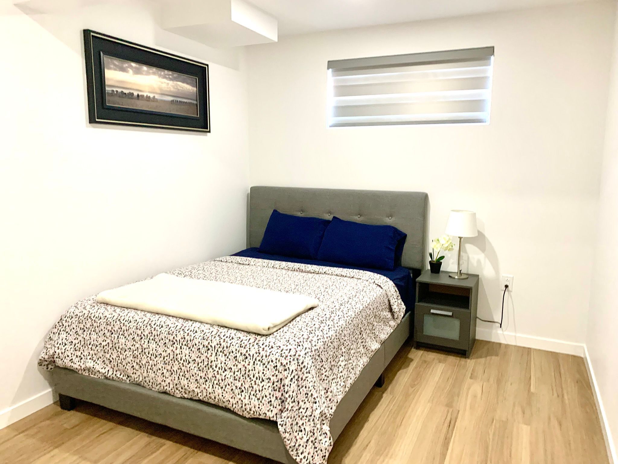 A neatly arranged bedroom with a bed, a bedside table with a lamp, a small potted plant, and a window with blinds.