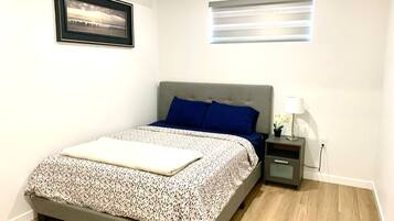 A neatly arranged bedroom with a bed, a bedside table with a lamp, a small potted plant, and a window with blinds.