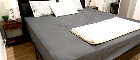 A neatly made bed with a grey bedspread and white pillows, a bedside table with a lamp, and a small plant.