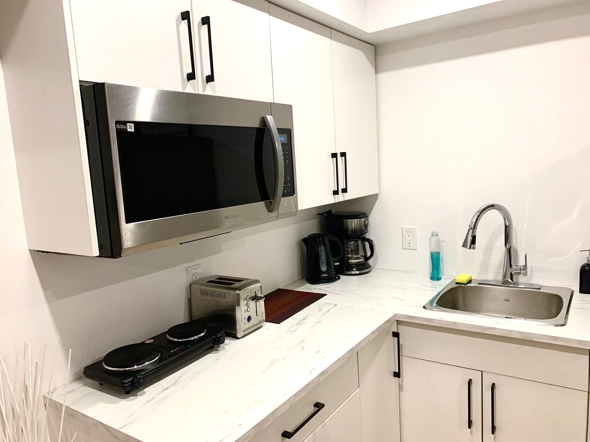 A modern kitchen with white cabinets, a stainless steel microwave, a coffee maker, a toaster, and a sink with a faucet.