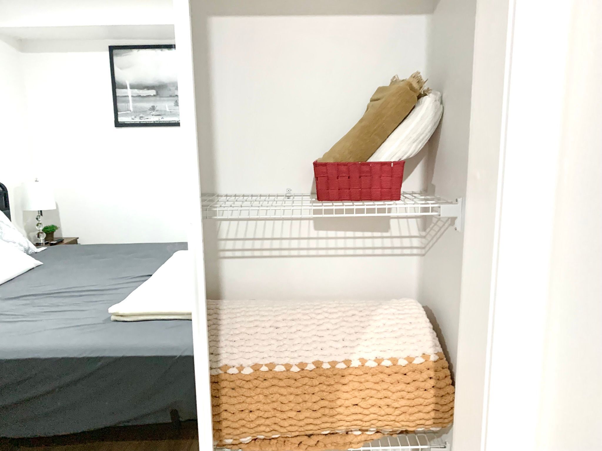 Une chambre à coucher avec un lit, une étagère sur laquelle se trouve un panier rouge, et un mur blanc orné d’un tableau encadré.