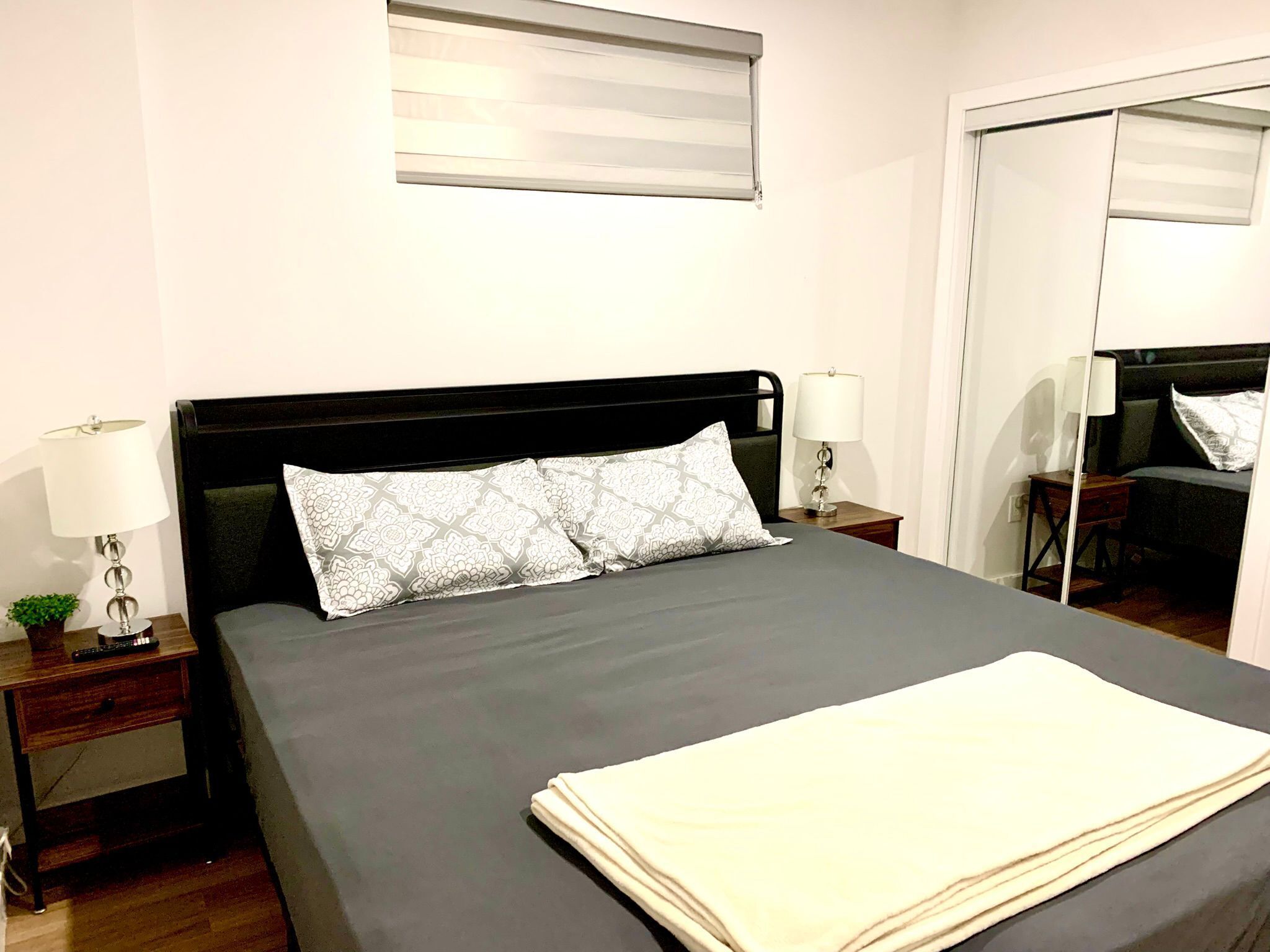 A neatly made bed with a dark headboard, two bedside lamps, and a small bedside table with a plant.