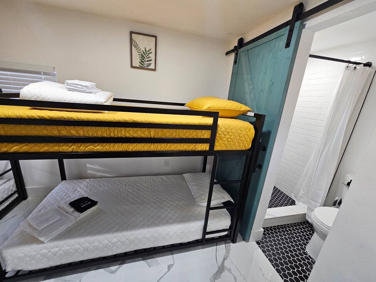 Habitación con literas, un colchón amarillo y uno blanco, una mesita de noche pequeña con un teléfono, y una puerta que da a un baño con inodoro y ducha blancos.