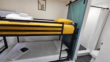 Habitación con literas, un colchón amarillo y uno blanco, una mesita de noche pequeña con un teléfono, y una puerta que da a un baño con inodoro y ducha blancos.