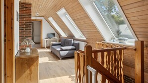 Ett sovrum på entresolplan med träribbor i taket, en grå soffa, ett skrivbord och ett fönster med utsikt över grönområden.