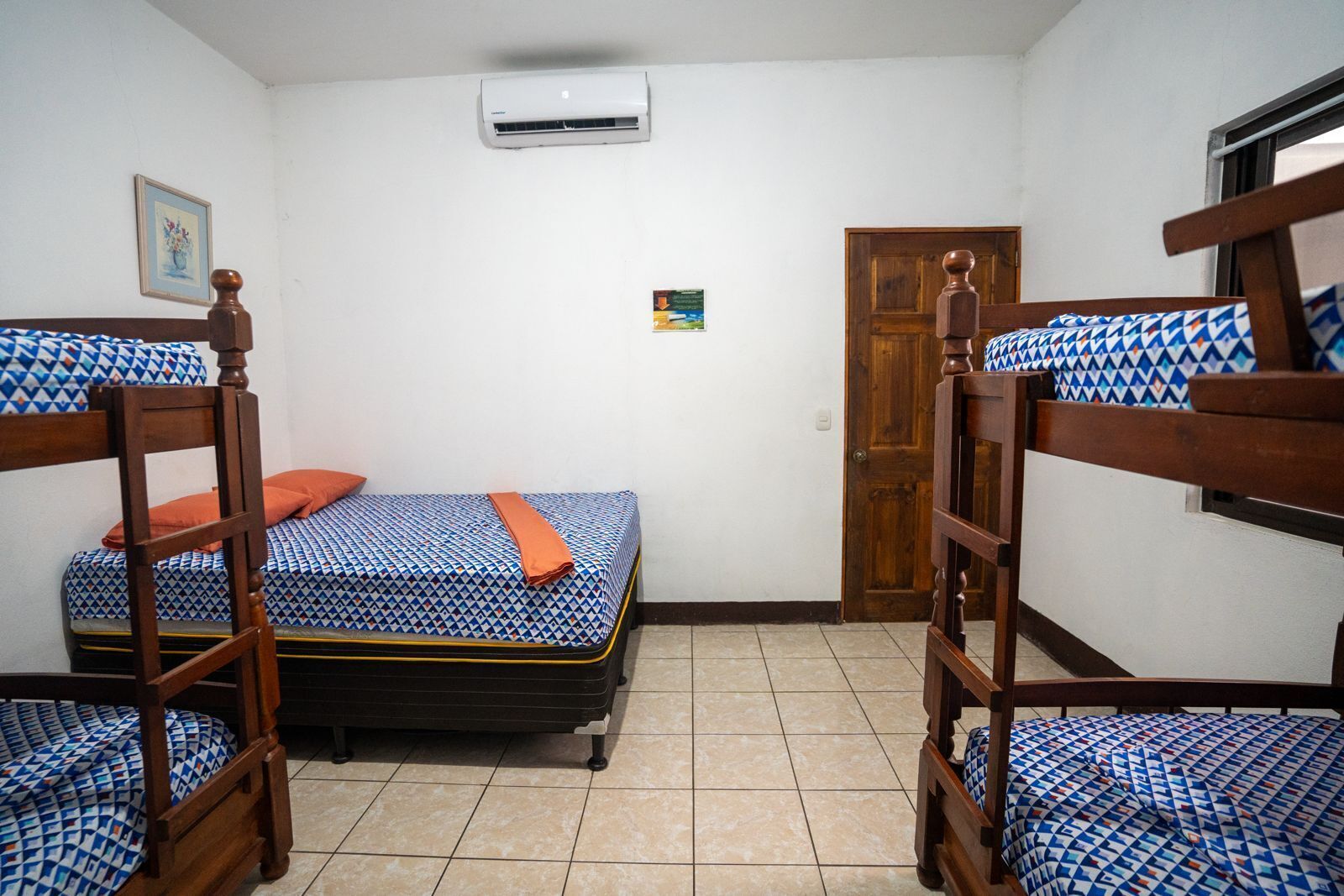 Een kamer met stapelbedden, een eenpersoonsbed, een deur, een aan de muur bevestigde airconditioner en een klein schilderij.