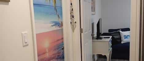 A hallway with a door leading to a bedroom. The bedroom has a bed, a desk, and a chair.
