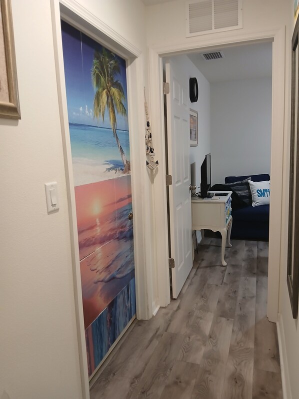 A hallway with a door leading to a bedroom. The bedroom has a bed, a desk, and a chair.