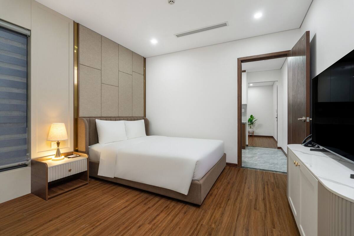 A modern hotel room with a large bed, a bedside table with a lamp, a flat-screen TV, and a wooden floor.