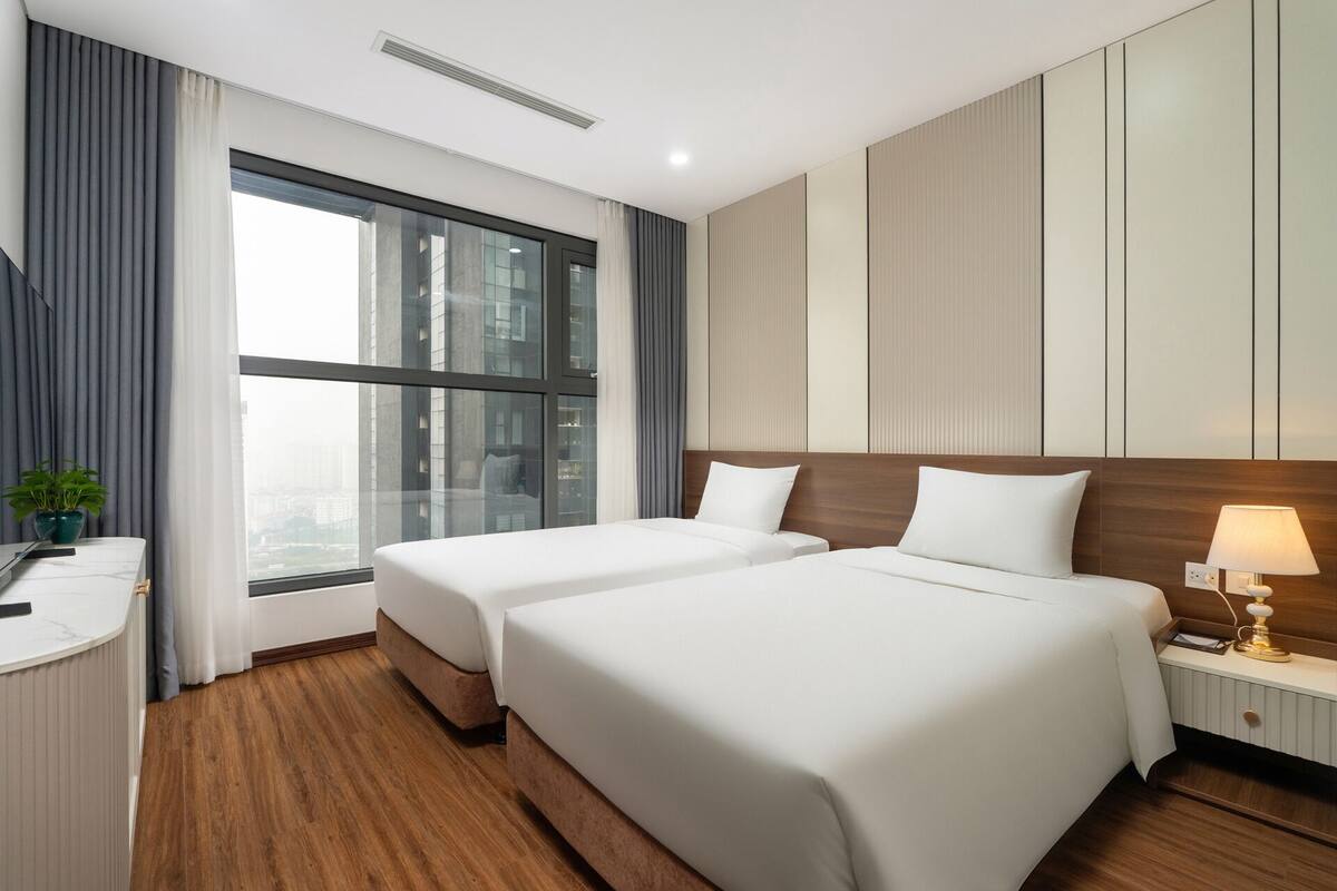 A hotel room with two beds, a desk, a lamp, and a view of the city through large windows.