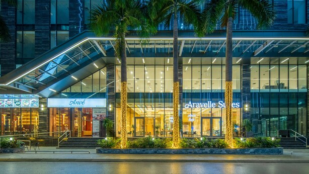 A modern hotel with a glass facade, palm trees, and illuminated signage. - ARAVELLE SUITES (Hanoi)
