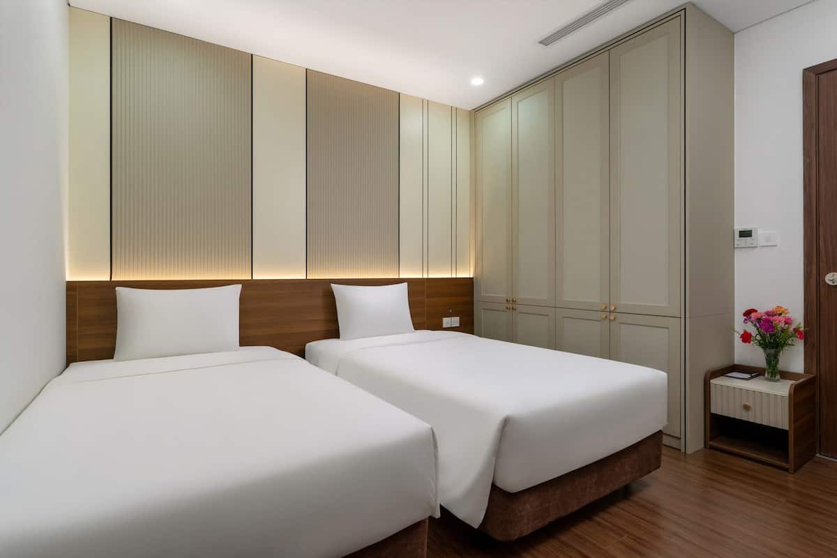 A hotel room with two beds, a wooden headboard, a large wardrobe, and a small bedside table with a vase of flowers.