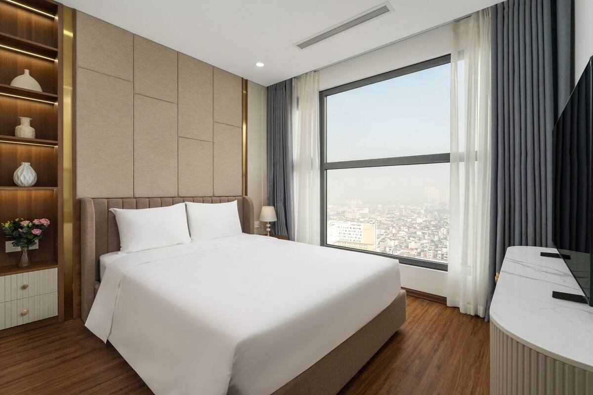 A hotel room with a large bed, a desk, and a view of a cityscape through a large window.