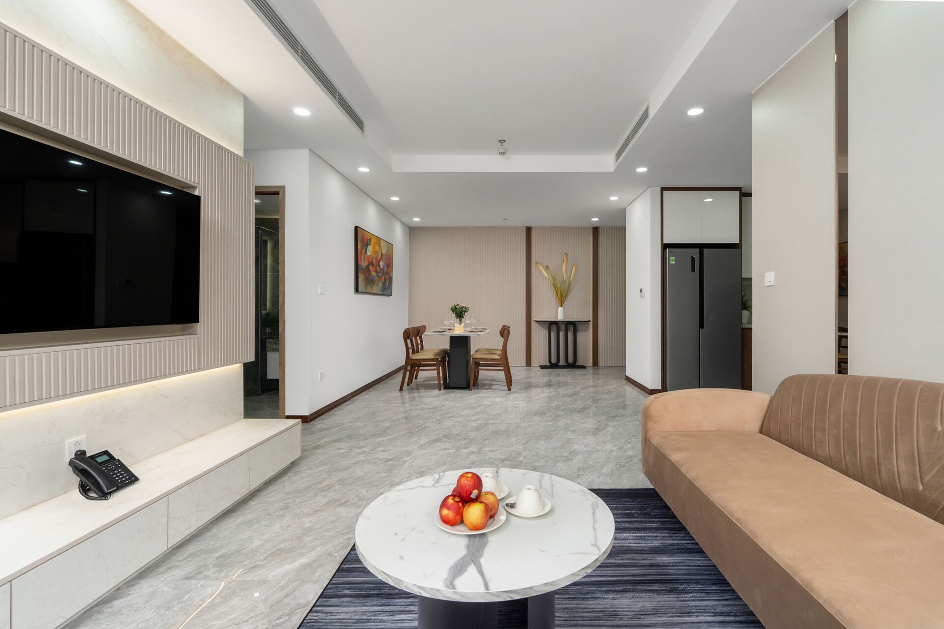 A modern living room with a flat-screen TV, a sofa, a coffee table with fruit, and a dining area with chairs and a table.