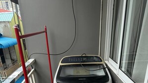 Una lavadora compacta y gris con etiqueta Samsung, ubicada sobre un piso de baldosas en un balcón junto a un tendedero de metal rojo.