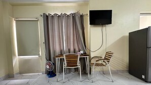 Una habitación con piso de baldosas, una mesa con dos sillas, un ventilador de pie, un televisor montado en la pared y un refrigerador.