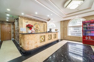 Uma área de recepção de hotel com balcão de mármore, mesa de atendimento, refrigerador de Coca-Cola e uma porta de madeira marcada com o número 002.