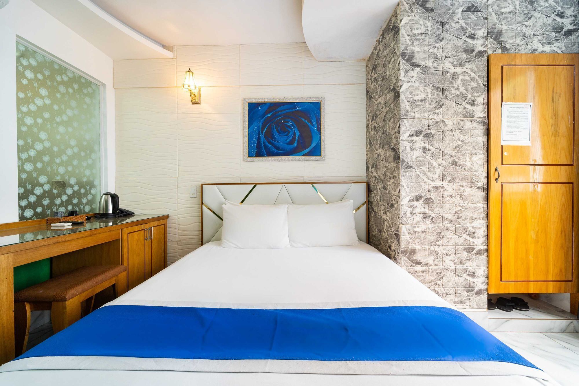 A hotel room with a bed, a wooden desk, a wall-mounted light, a framed abstract painting, and a stone-textured wall.