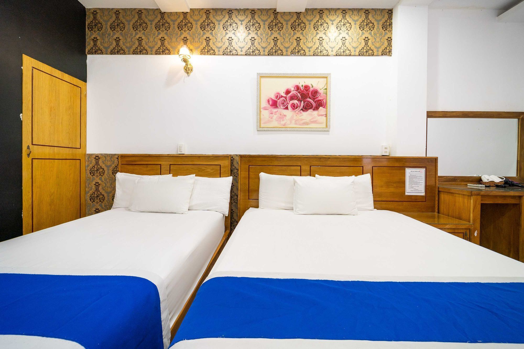 A hotel room with two beds, a wooden wardrobe, a desk with a mirror, and a wall-mounted light fixture.