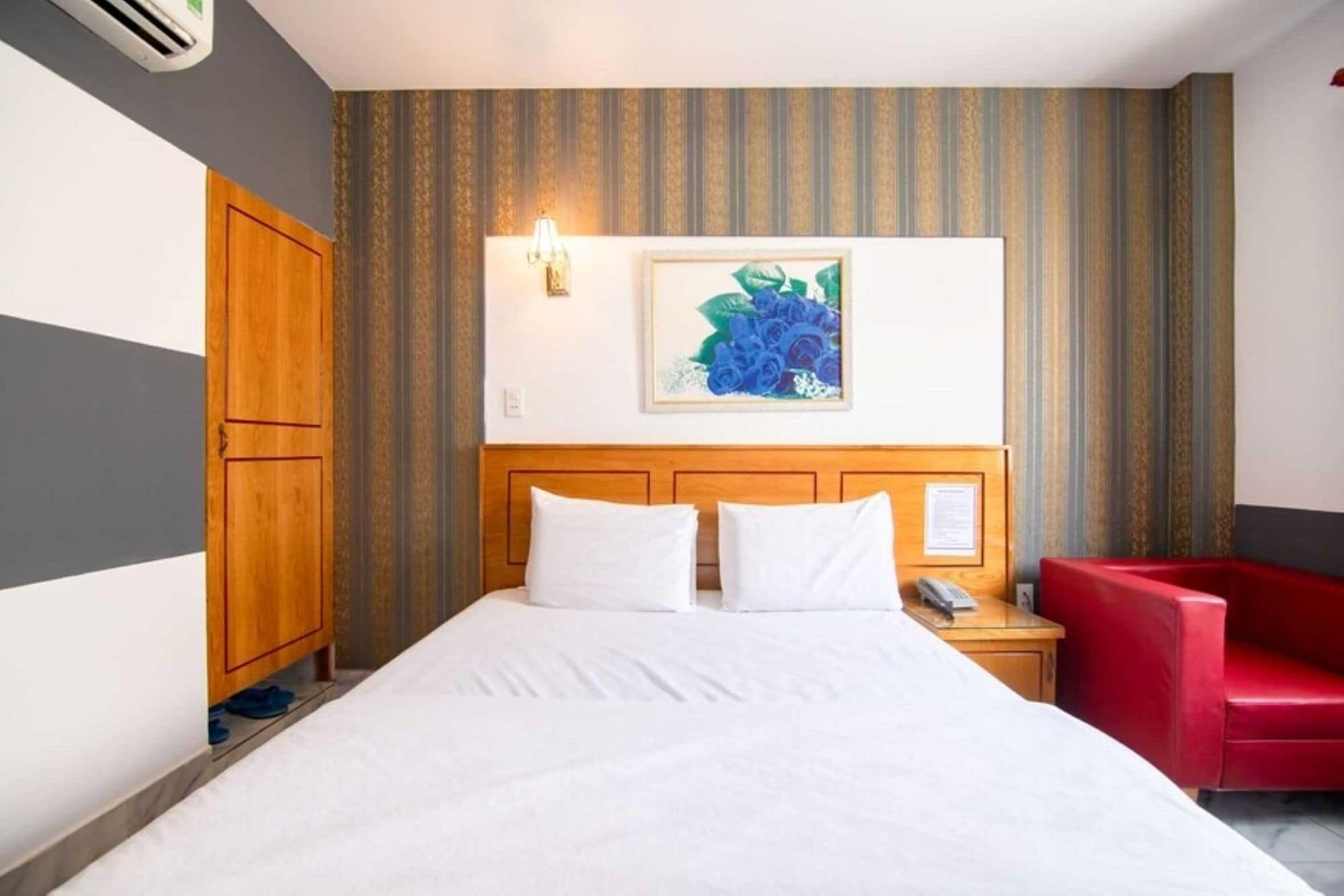 A hotel room with a bed, a wooden wardrobe, a red chair, and a wall-mounted artwork.